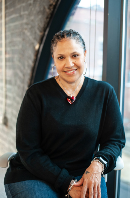 Artist and educator Liz Ivey wearing a black sweater with a red necklace, sitting indoors against a window and brick background.