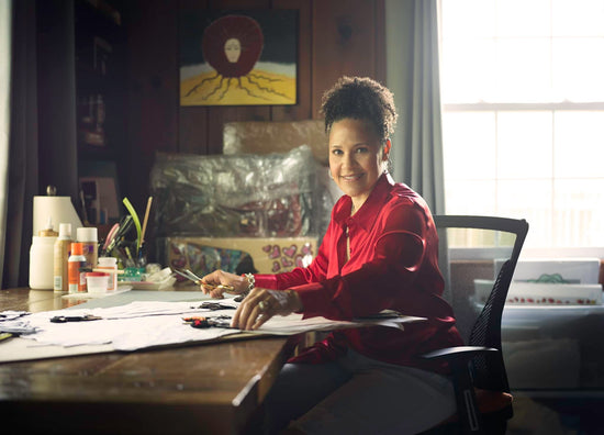 Artist Elizabeth Ivey sitting at a desk wearing a red shirt in a room with art supplies and a painting on the wall.